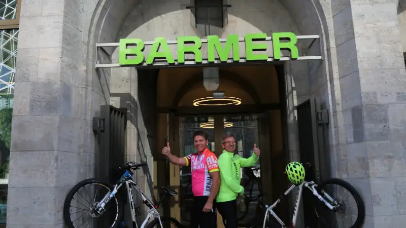 Zwei Barmer Mitarbeiter, die mit ihren Fahrräder ein V in der Eingangspforte von Barmer bilden