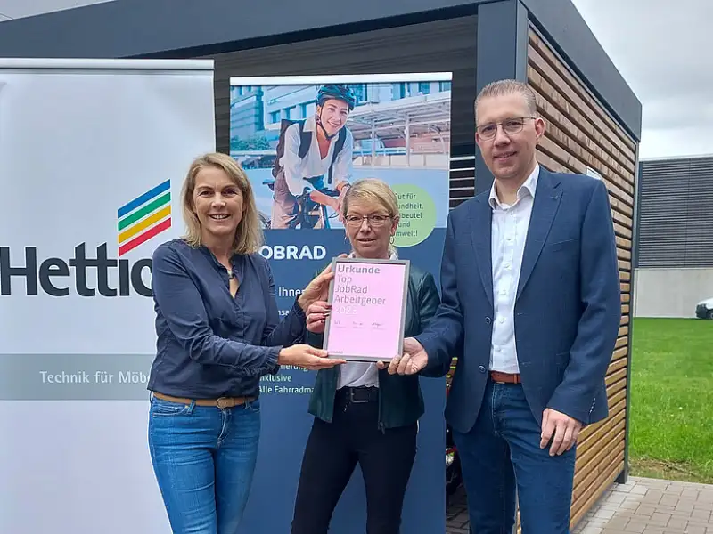 Monika Titze, von der Hettich Gruppe, steht zusammen mit einer Frau und einem Mann vor einem Poster mit dem Schriftzug Hettich und dem Hettich Logo. Sie alle tragen blaue Geschäftskleidung